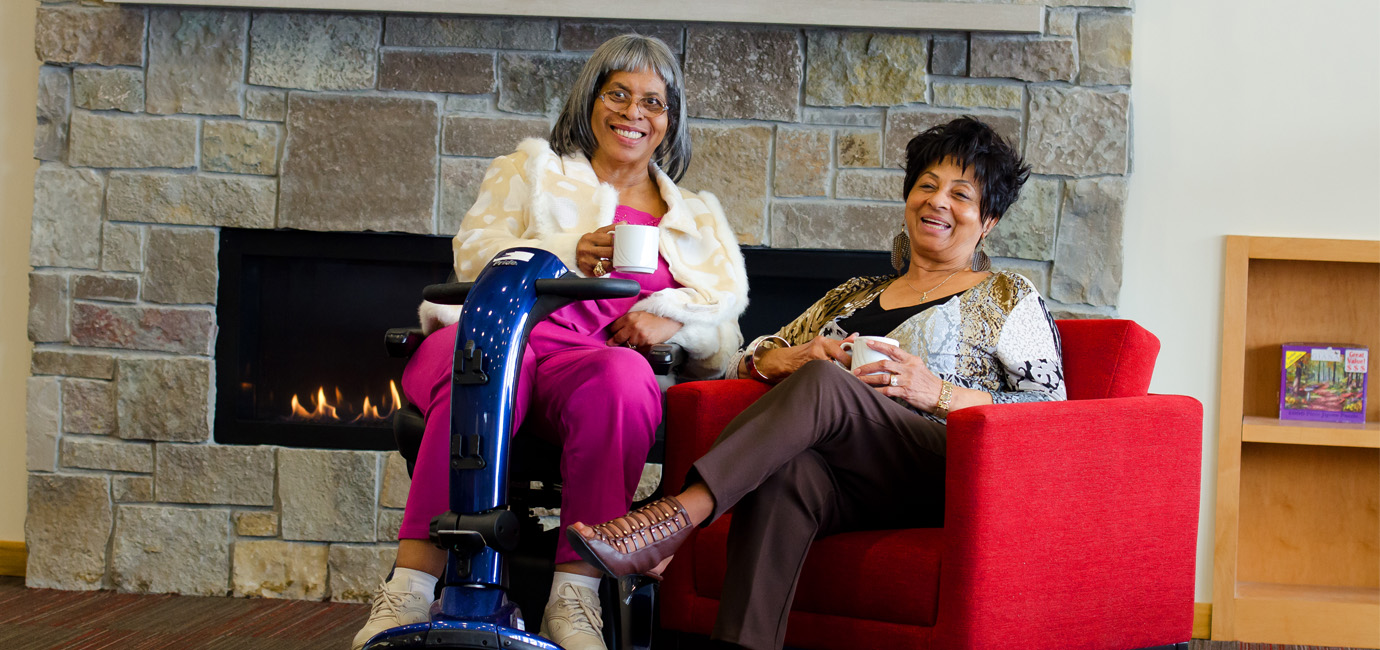 Two women enjoying coffee by a fireplace, one seated in a red chair and the other on a mobility scooter.